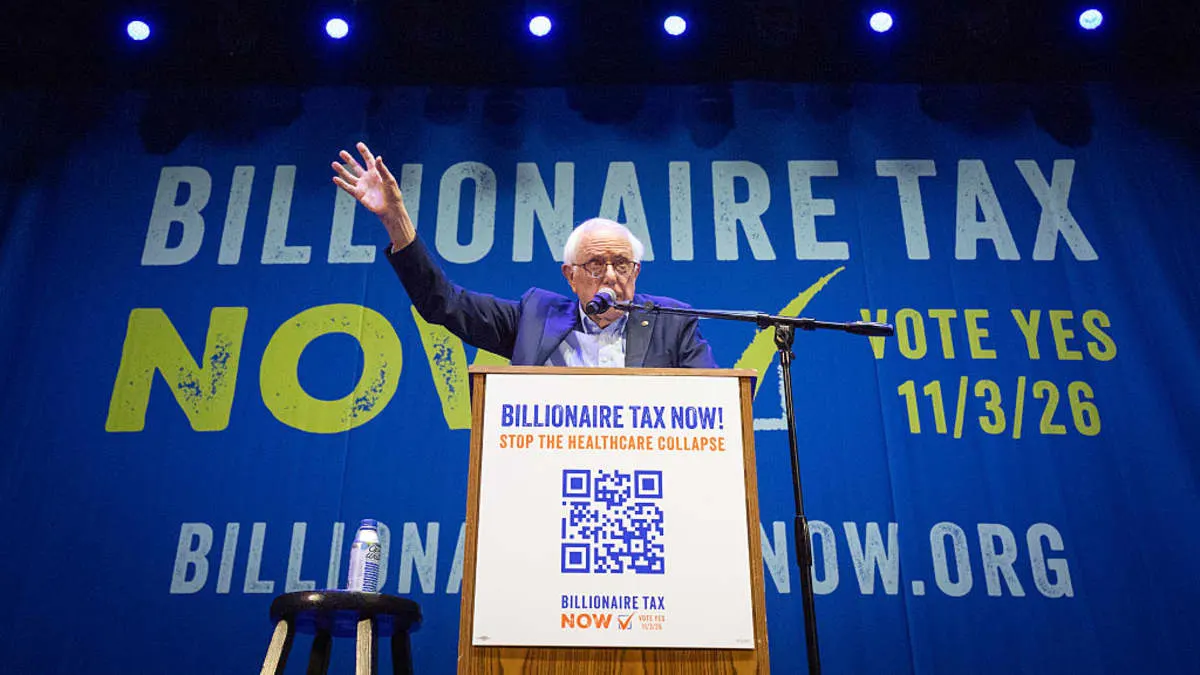 Older man at podium raising hand beneath blue 'Billionaire Tax Now' campaign banner, vote yes 11/3/26.