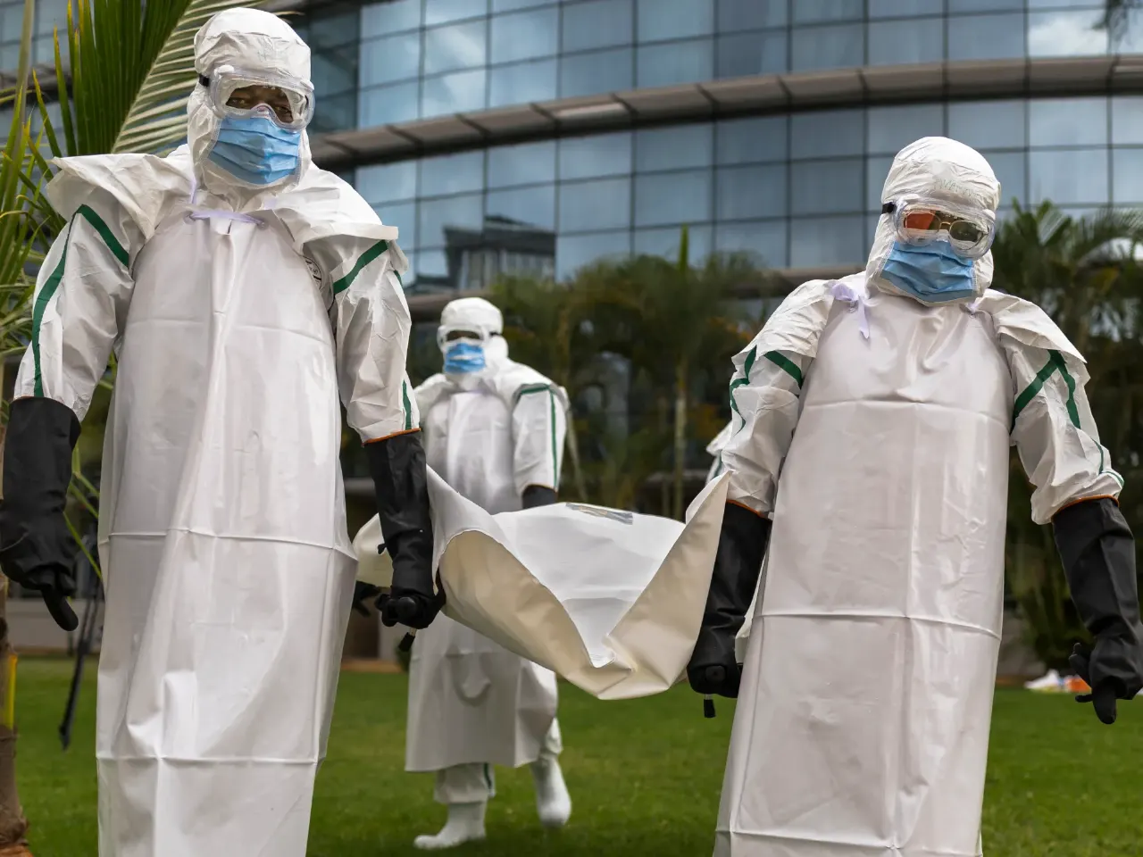 Workers in full white protective suits carry a stretcher across a lawn outside a glass building.