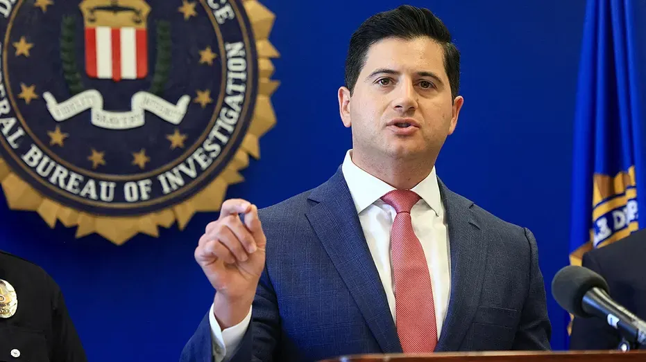 Man in suit and red tie gestures while speaking at podium before FBI seal and flag