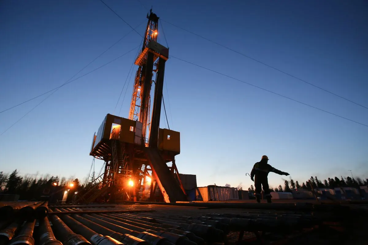 Oil drilling rig silhouetted at dusk with a worker gesturing beside pipes.