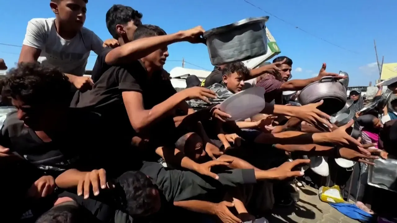 Crowd of people reaching out with metal cooking pots, pressing forward to receive food aid outdoors.