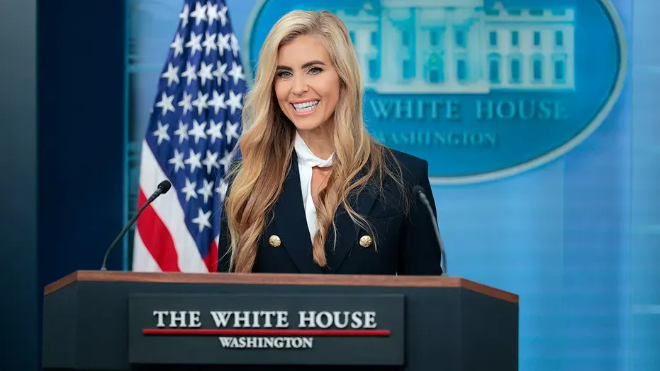 Smiling woman with long blonde hair at a White House podium beside an American flag.