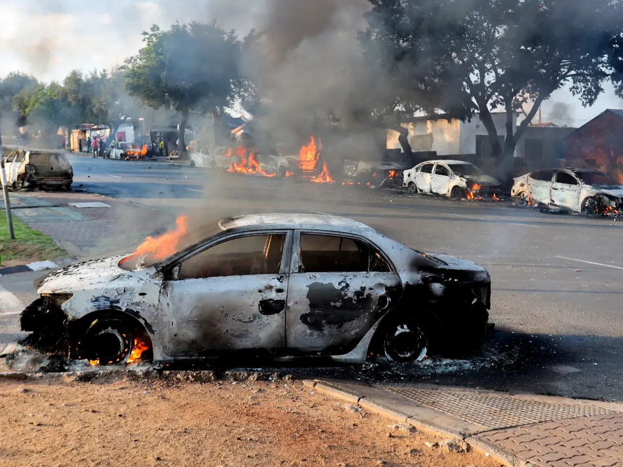 Burnt-out cars on a street with smoke and fire, showing aftermath of unrest