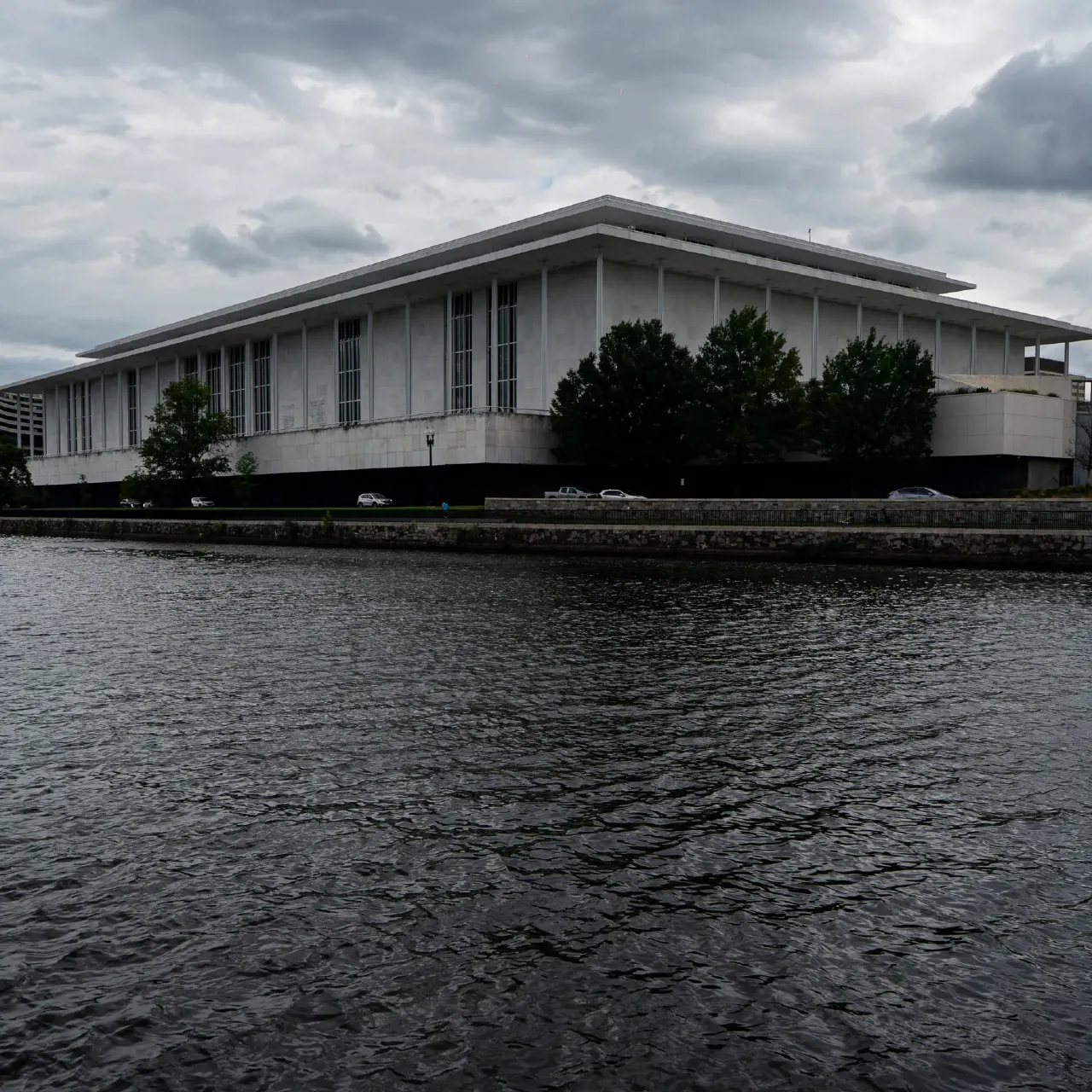 Concrete modernist building with flat roof beside a river under cloudy sky.