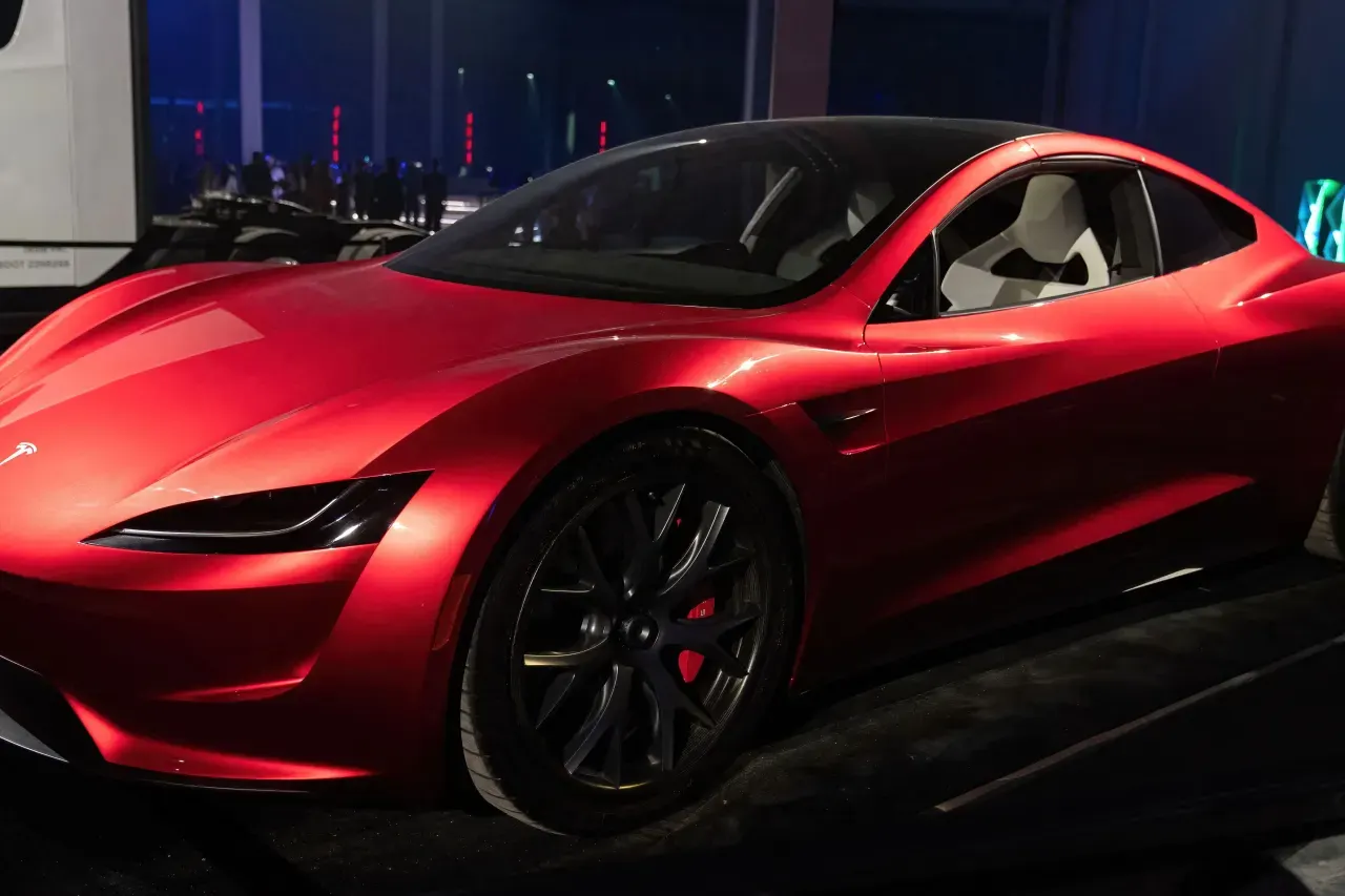 Red sports car with black wheels displayed on dark stage, Tesla badge visible.