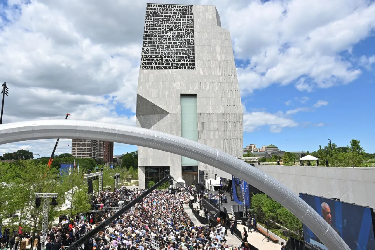 Large crowd gathered before a modern stone tower with a white curved arch, at a public ceremony.