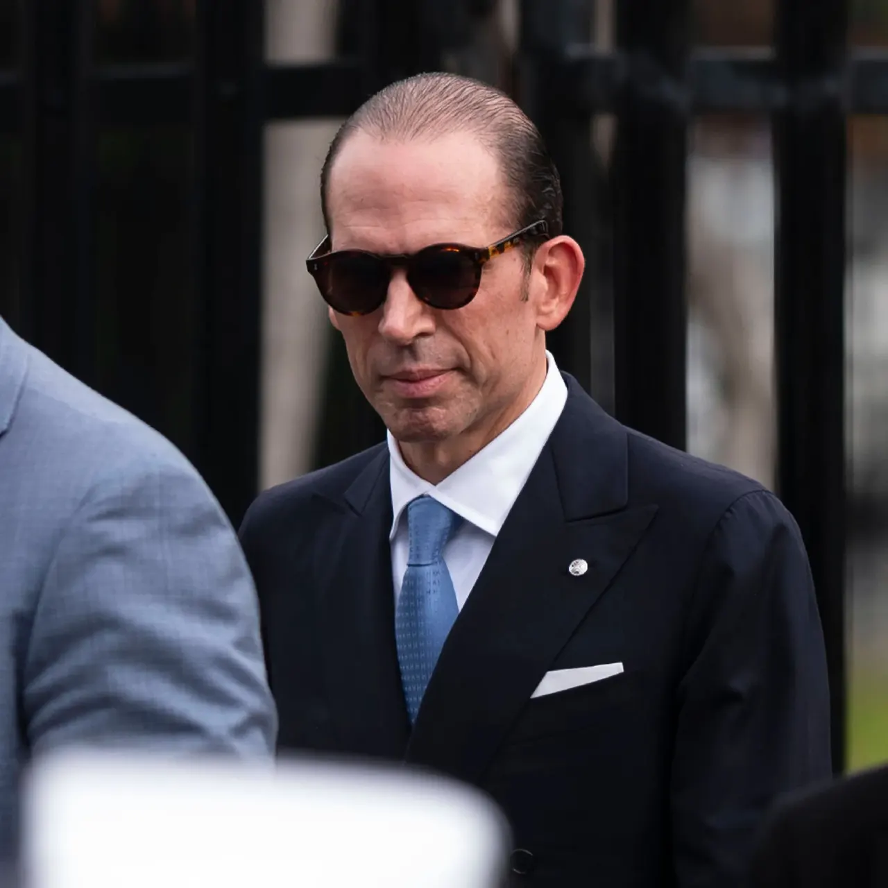 Man in dark suit, blue tie and tinted sunglasses standing outdoors near black iron gate.