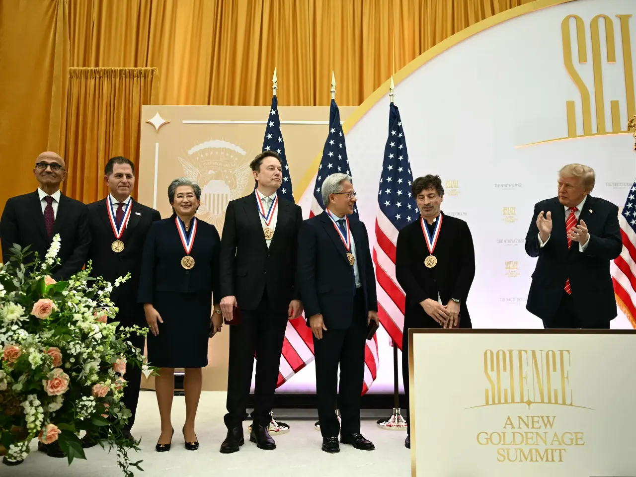 Five medal-wearing executives stand on stage at a Science Summit as a man applauds nearby.