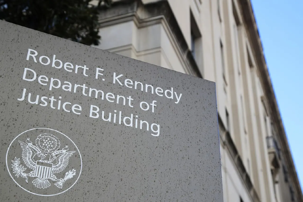 Stone sign with Department of Justice seal beside a stately building facade under blue sky.
