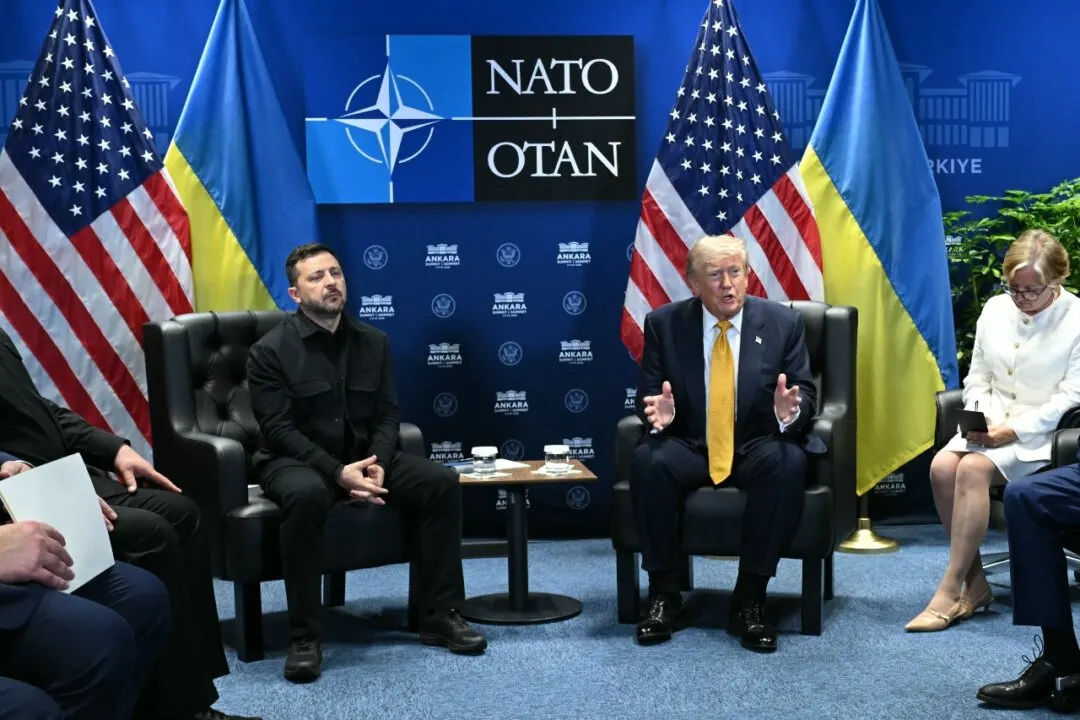 Two men seated in armchairs at a NATO summit, flanked by US and Ukrainian flags, talking.