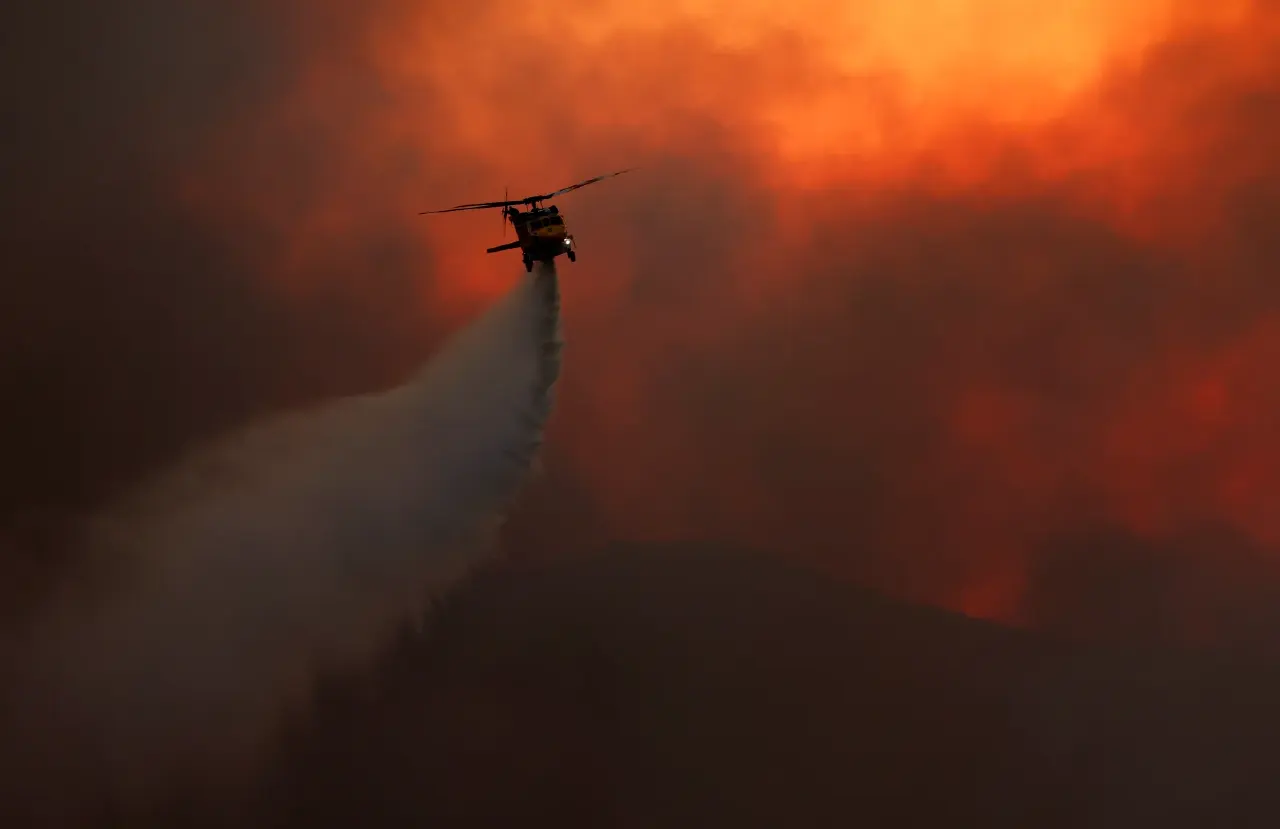 Helicopter dropping water on a wildfire against an orange, smoky sky at dusk.
