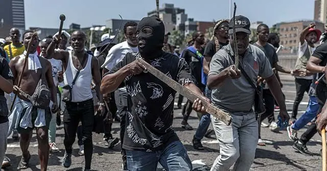 Masked man with wooden stick leads crowd of men marching down a city road