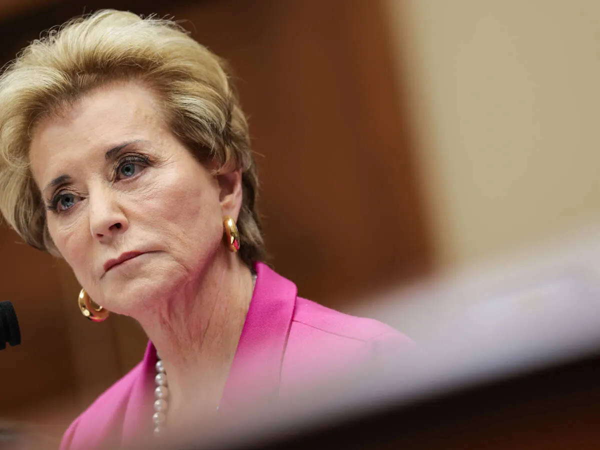 Older woman with blonde hair in pink jacket, looking serious, at a microphone in a hearing room.