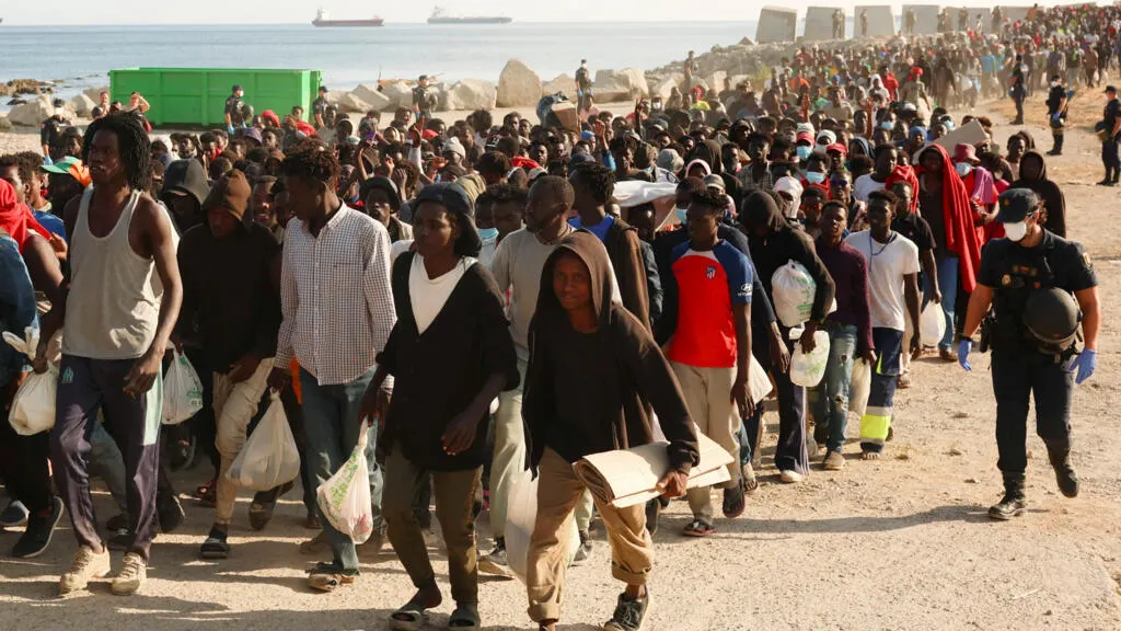 Large crowd of migrants walking along a rocky shoreline, carrying bags, escorted by a uniformed officer.