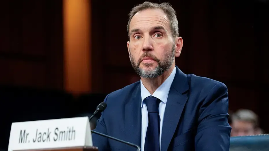 Bearded man in navy suit at hearing table with nameplate and microphone.