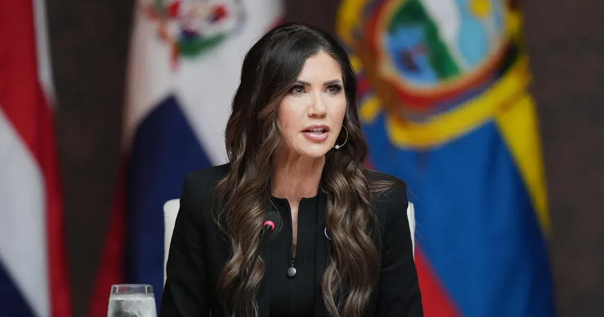 Woman with long wavy hair speaking at a microphone before several national flags.