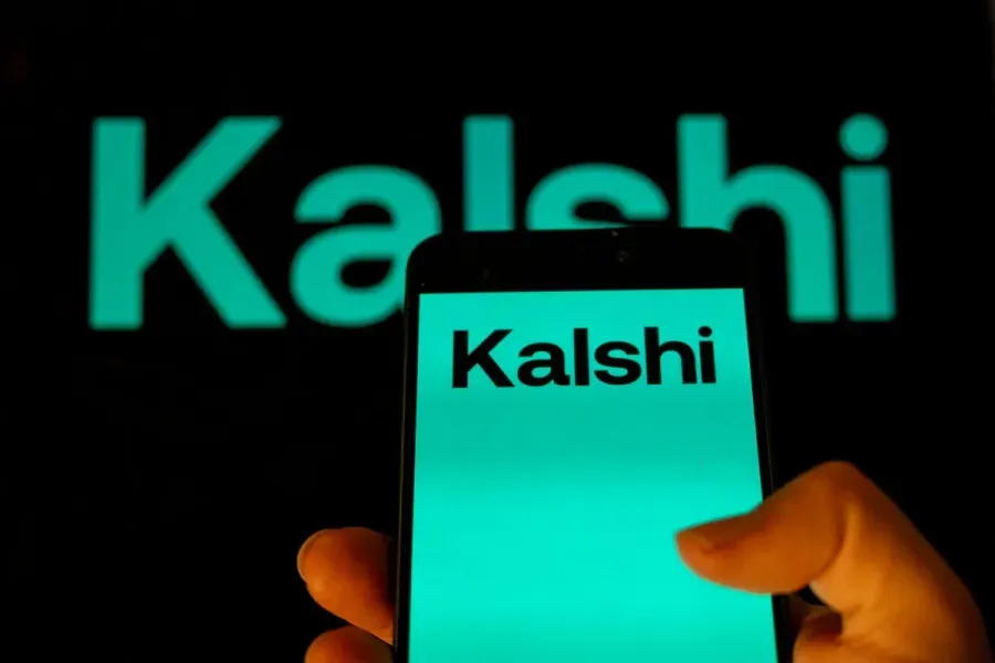 Hand holding smartphone displaying Kalshi logo, with large Kalshi logo glowing behind.