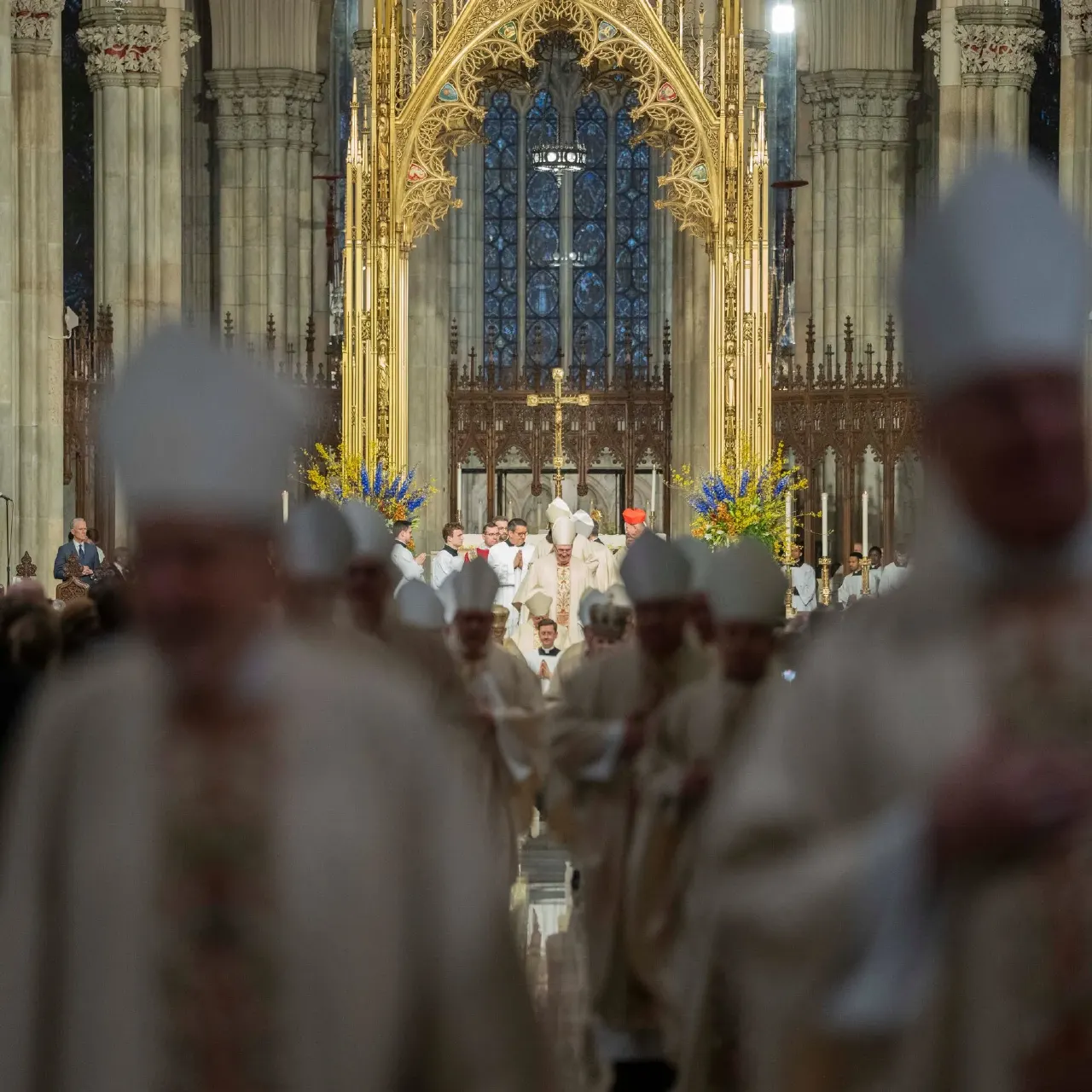Blurred mitred clergy in foreground processing toward altar inside a Gothic cathedral.