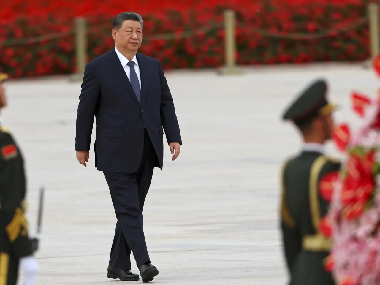A man in dark suit walks past uniformed honour guards beside red flowers on a plaza.