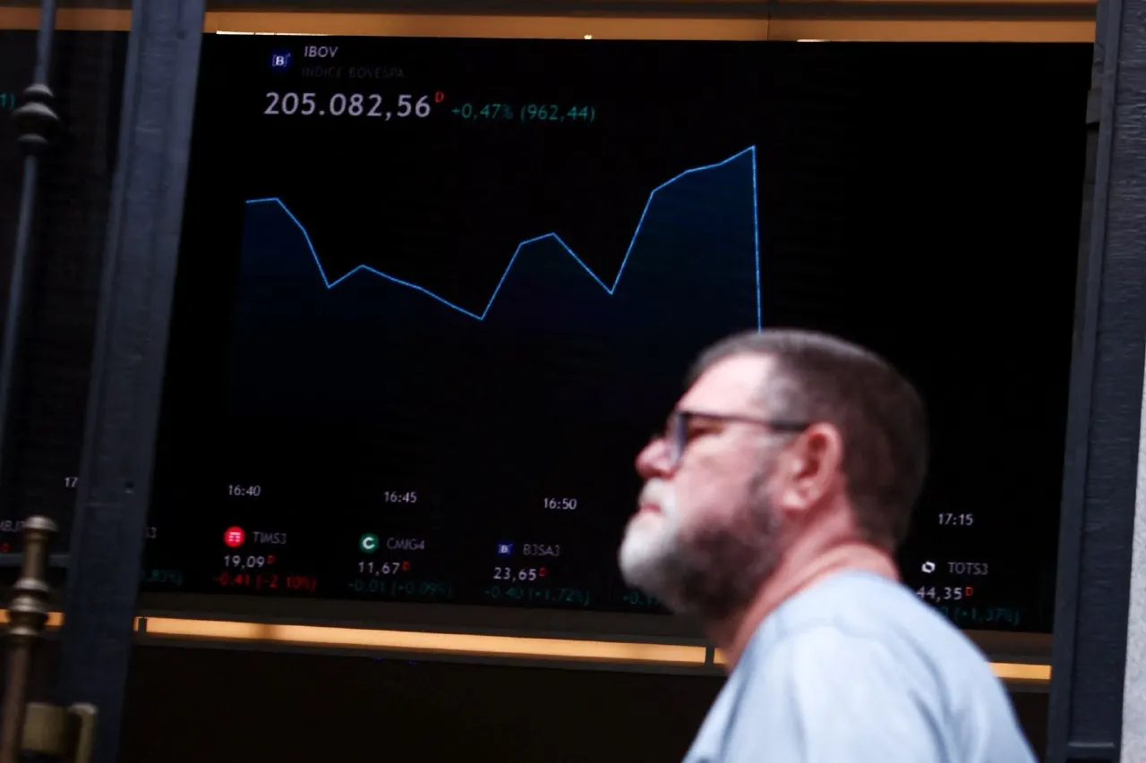 Man in glasses walks past a screen showing a downward-trending Ibovespa stock index chart.