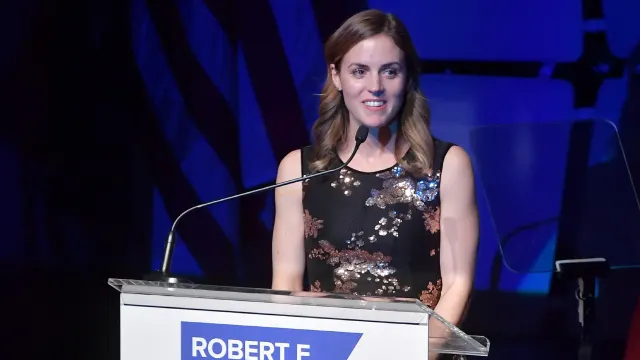 A smiling woman in a sparkly black dress speaks at a podium with a blue 'ROBERT F' sign.