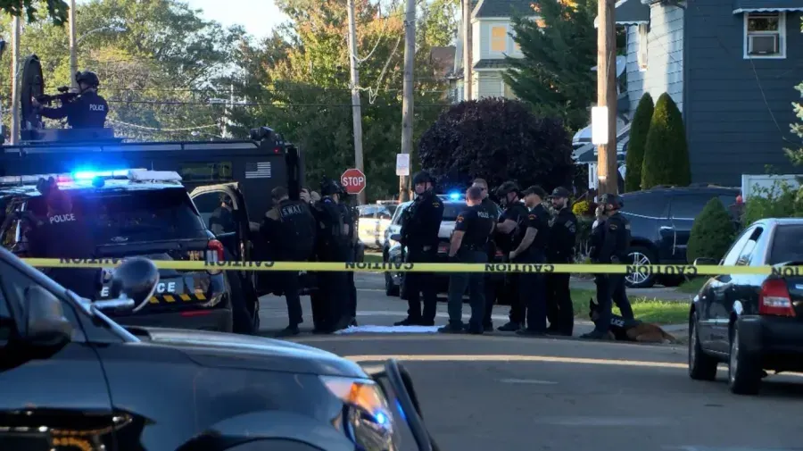 Police in tactical gear and SWAT vehicles behind yellow caution tape on a residential street.