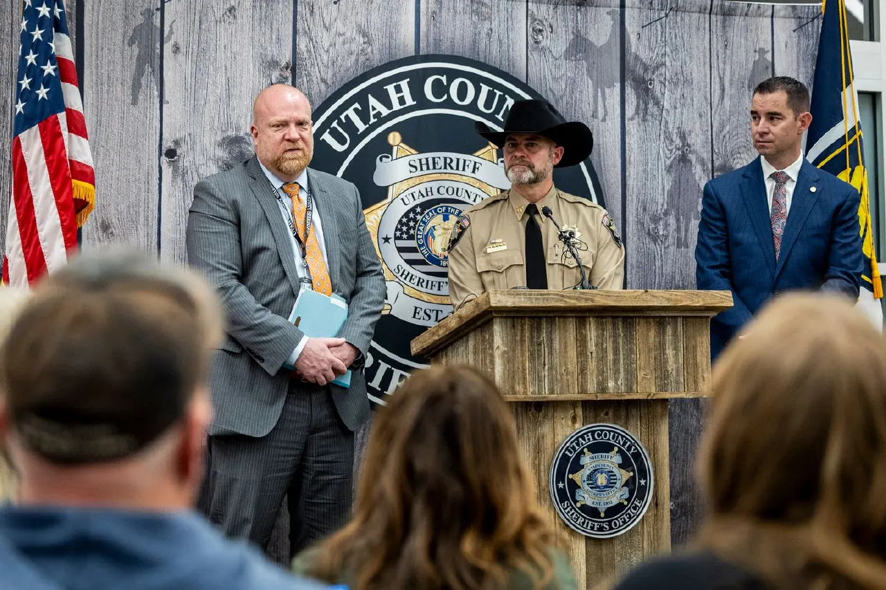 Uniformed sheriff speaks at podium before Utah County Sheriff's Office seal, flags, and audience.