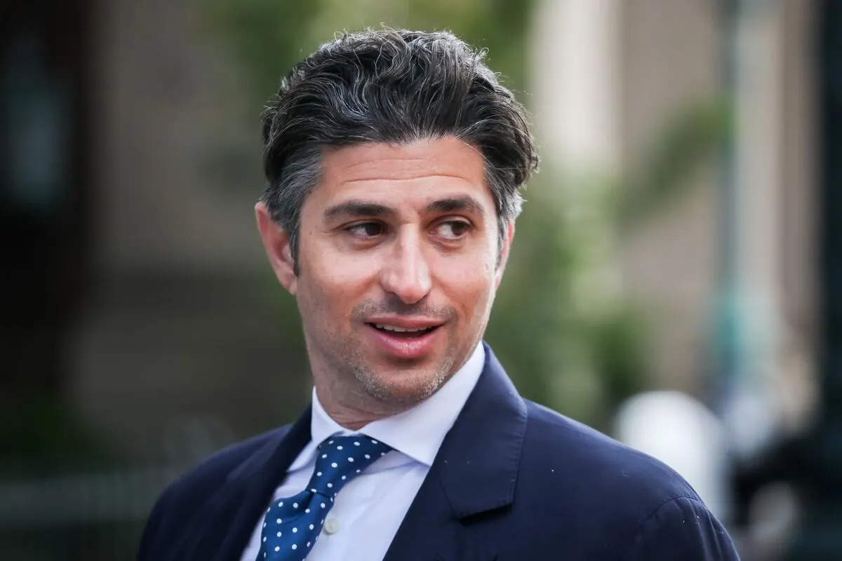 Man in navy suit, white shirt and polka-dot tie, looking to the side outdoors.