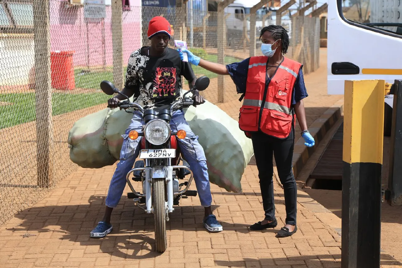 Health worker in red vest and mask checks a motorcyclist's temperature at a gated entrance.