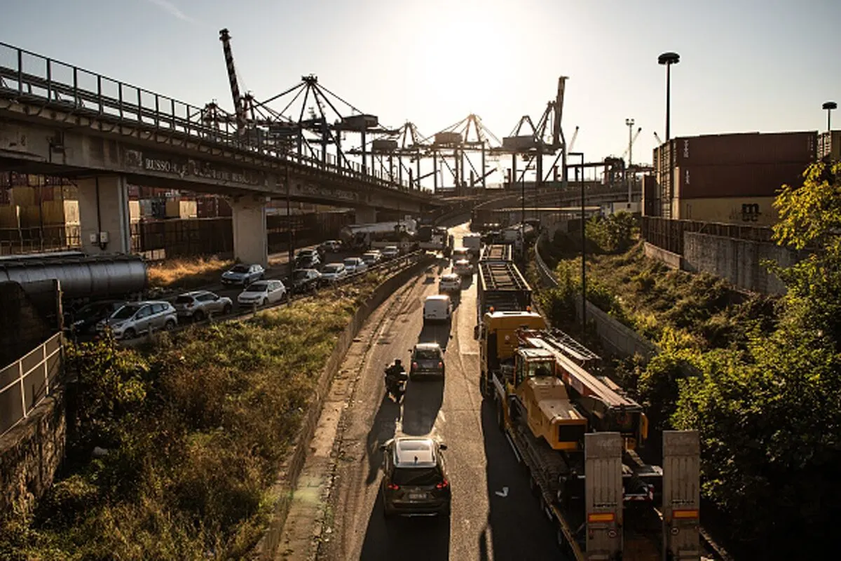 Congested highway with cars and trucks beside a port with cranes at sunset.