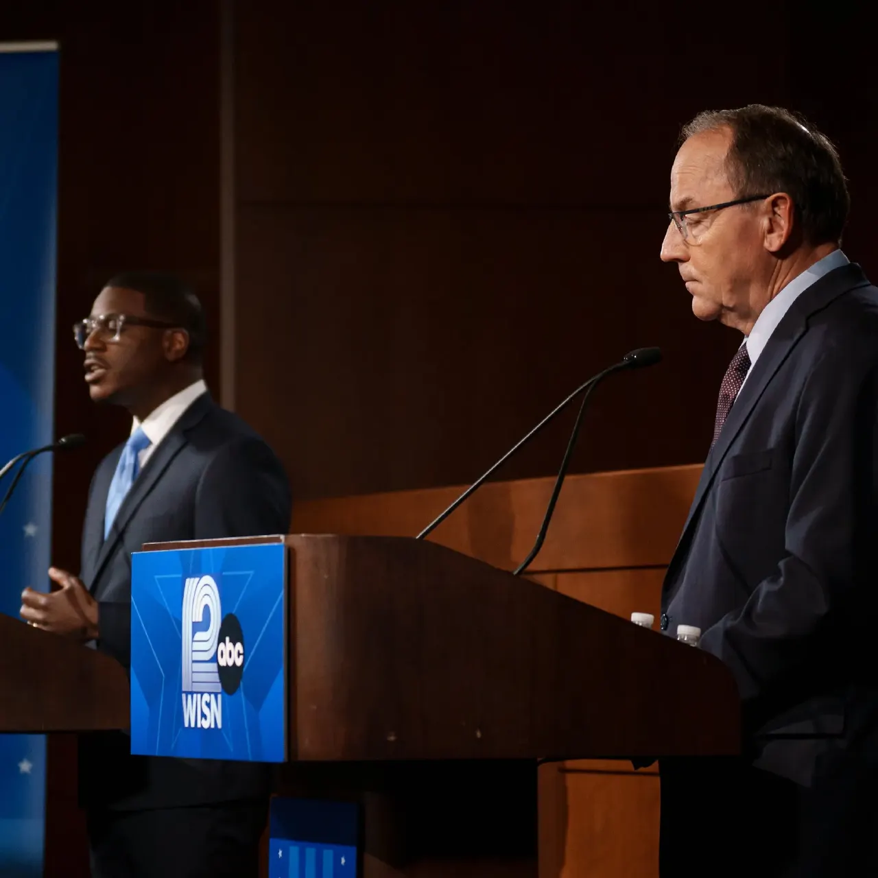 Two men in suits at wooden debate podiums with microphones, one speaking, one looking down.