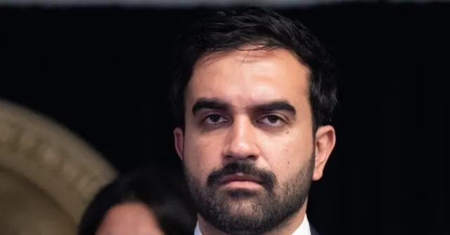 Close-up of a bearded man with dark hair in a dark setting, looking forward seriously.