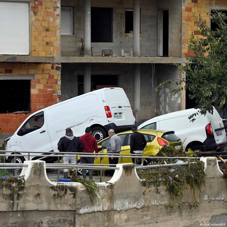 Men stand by a flooded street, looking at cars piled up and damaged by floodwater.
