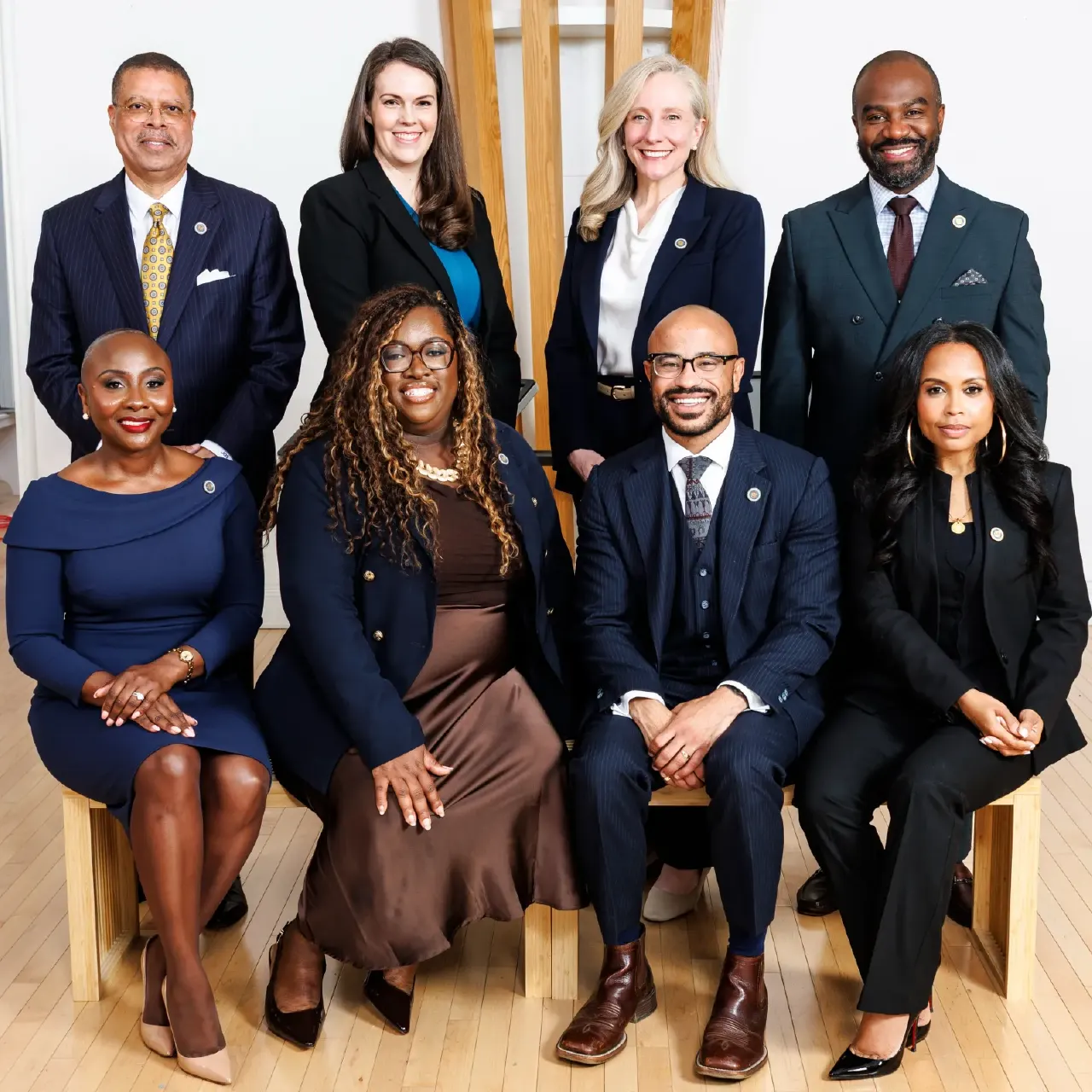Group portrait of eight smiling professionals in suits posed in a bright studio setting.
