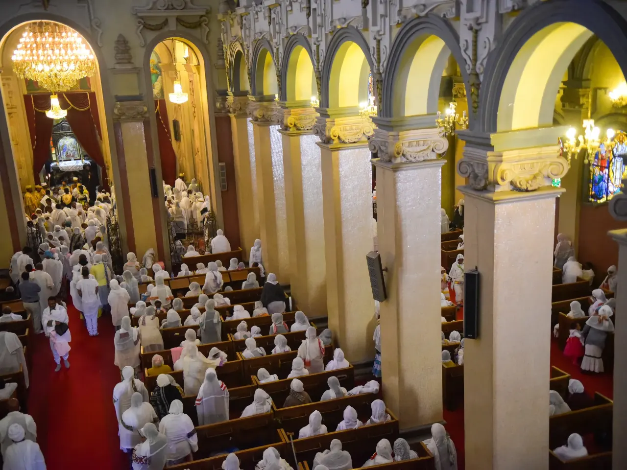 Worshippers in white robes fill pews of an arched church interior with red carpet.