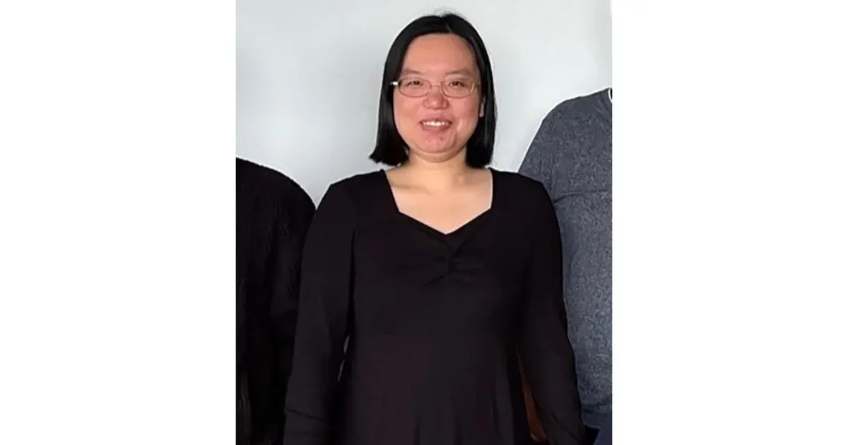 Smiling woman with glasses and shoulder-length dark hair in black top, against plain background.