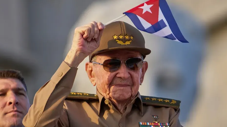 Elderly man in military uniform and cap raising a fist while holding a Cuban flag, wearing sunglasses