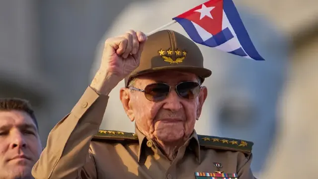 Elderly man in military uniform and cap raising a fist while holding a Cuban flag, wearing sunglasses