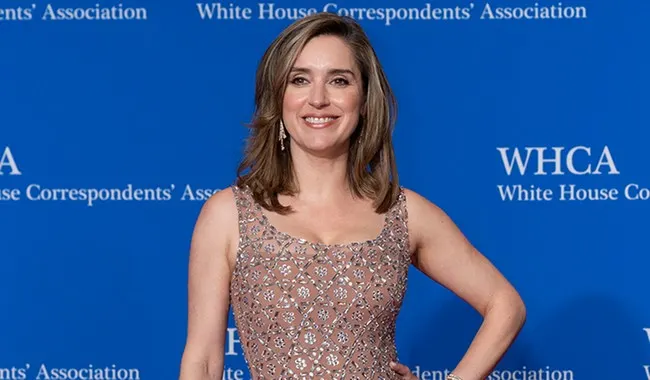 Smiling woman in embellished dress posing before blue White House Correspondents' Association backdrop.