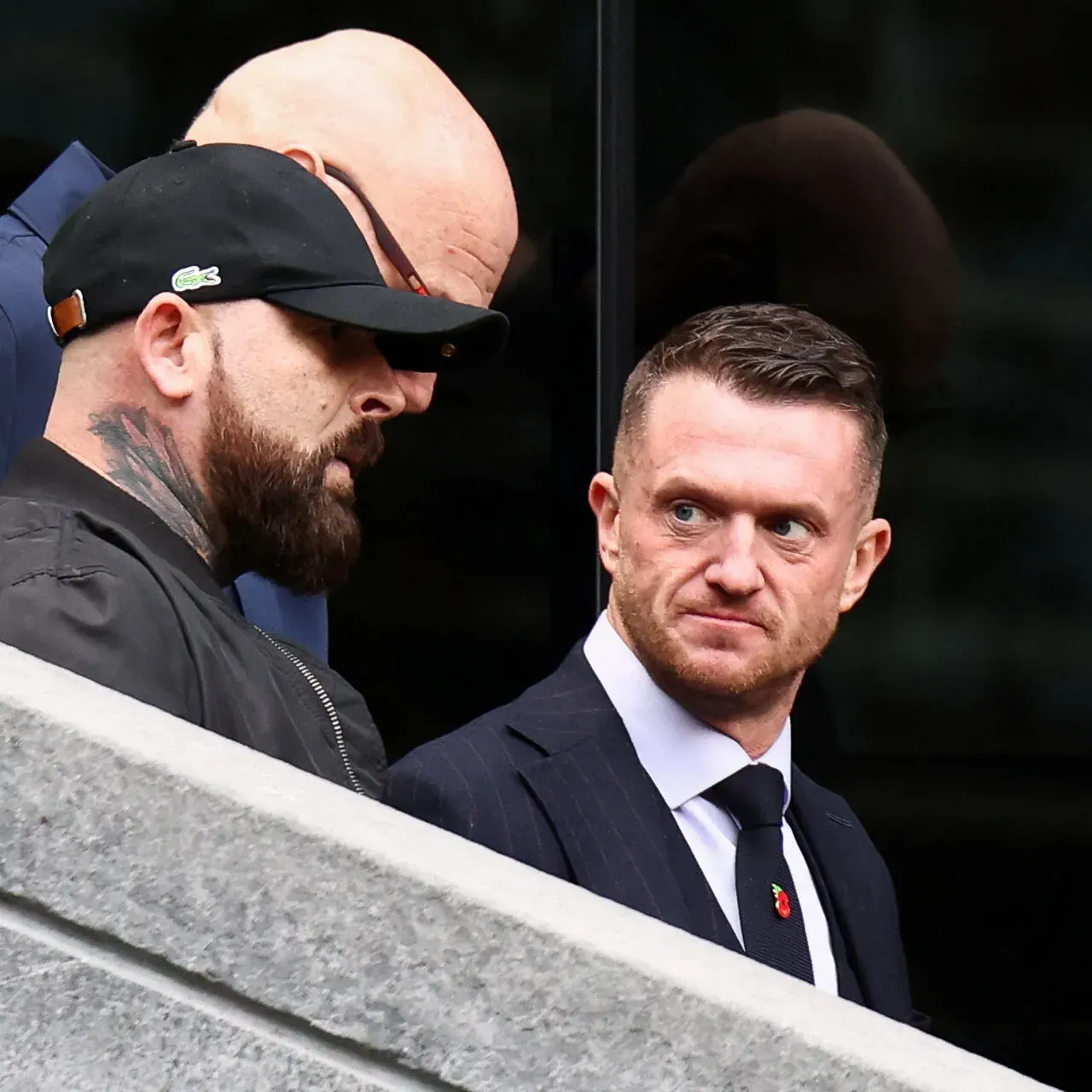 Two men in dark clothing on a stone staircase; one in a cap, one in a suit glancing sideways.