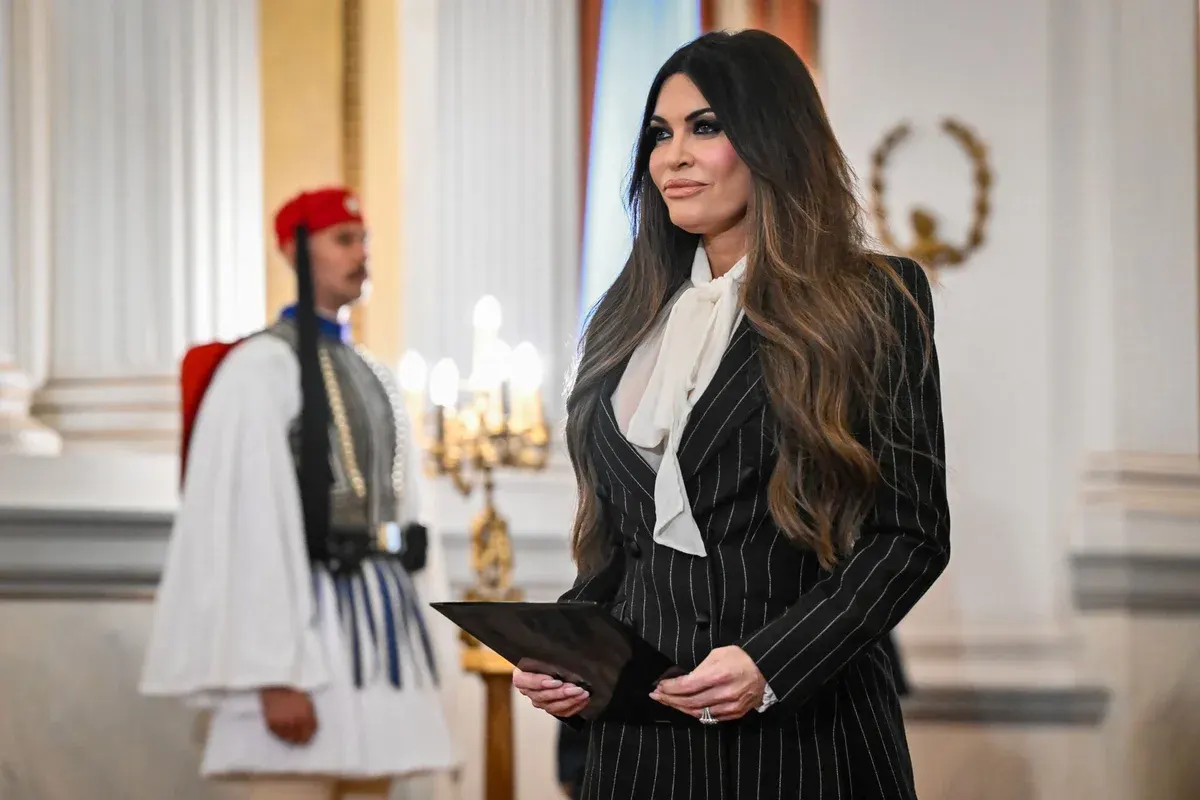 Woman in pinstripe suit holding a folder, standing near a uniformed guard in a formal, ornate hall.