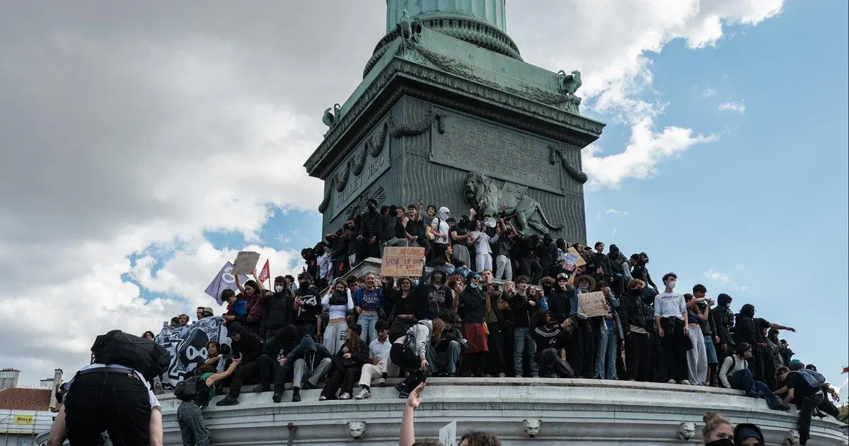 Large crowd of young protesters sitting and standing atop a green monument at Bastille Square, holding signs.