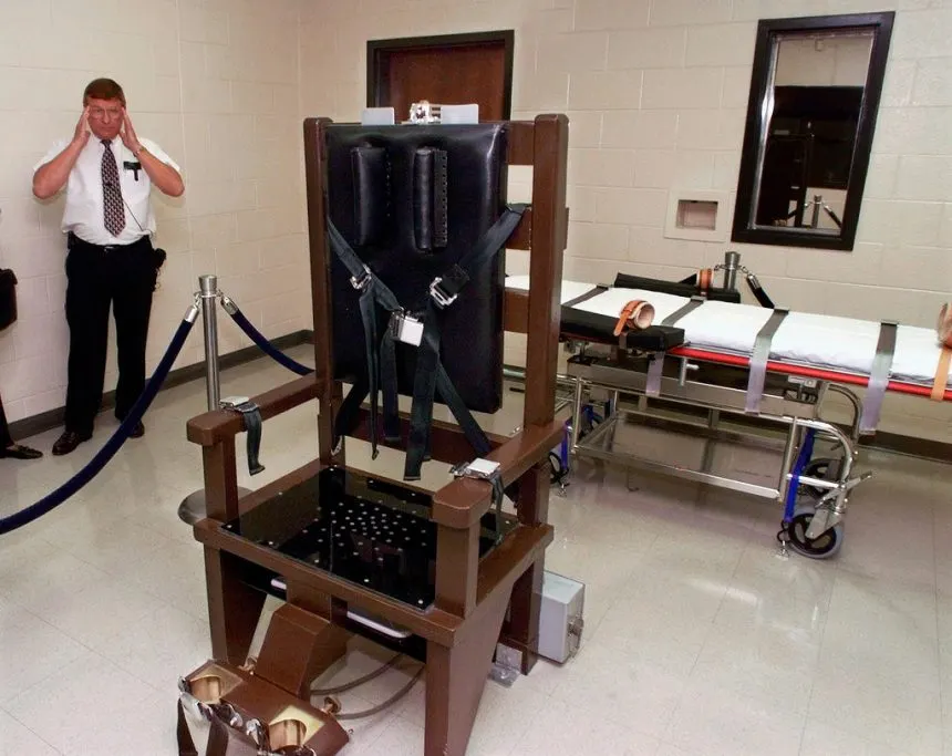 Execution chamber with electric chair and gurney, with a uniformed official standing nearby.