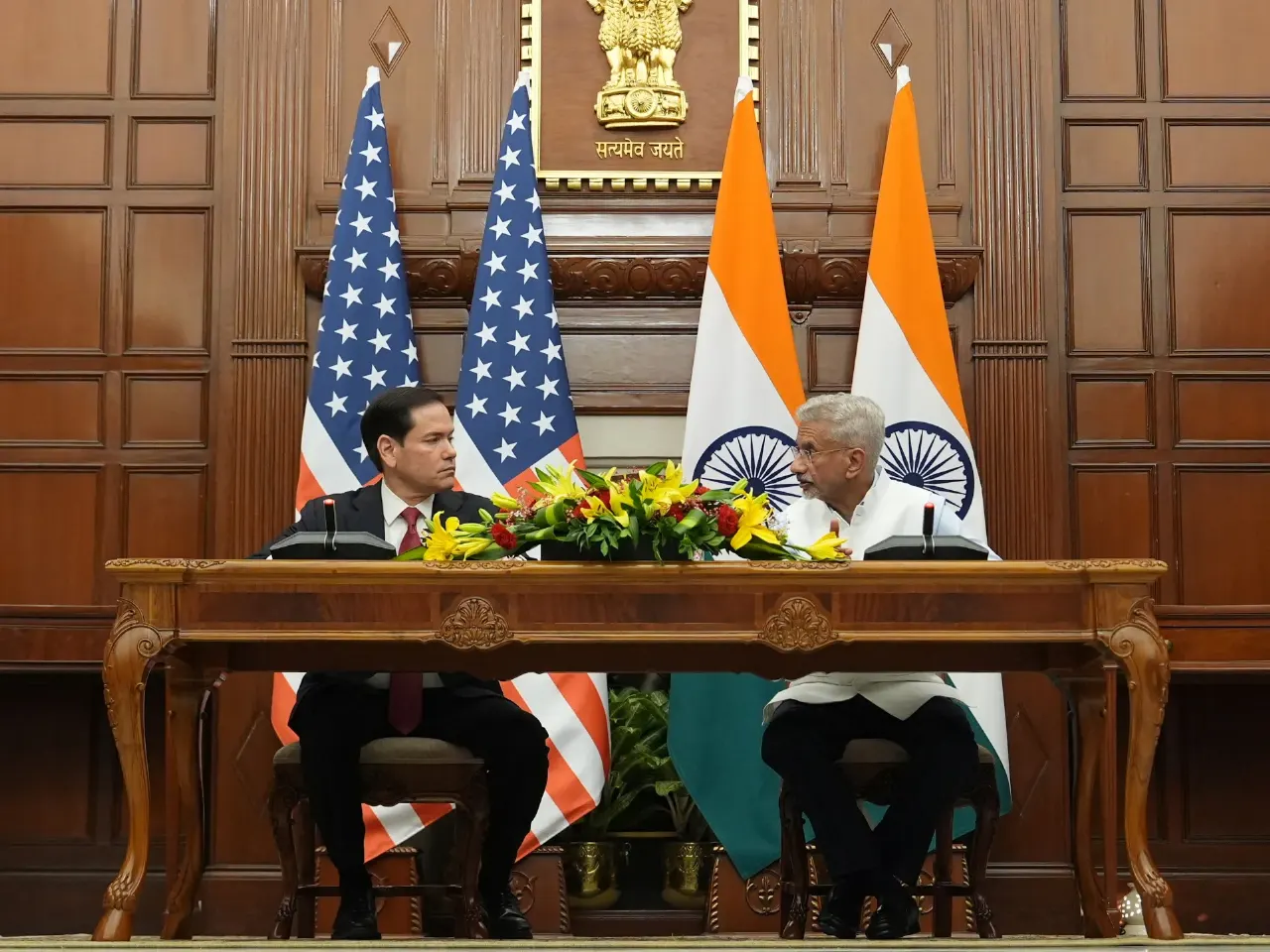 Two officials seated at a formal table before US and Indian flags, with flowers and an emblem.