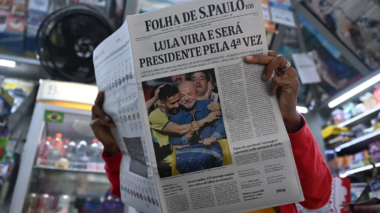 Person in red sweater holds Folha de S.Paulo newspaper with Lula victory headline in a shop.