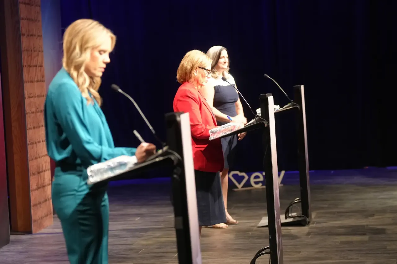 Three women stand at debate podiums on a stage with microphones, one writing notes.