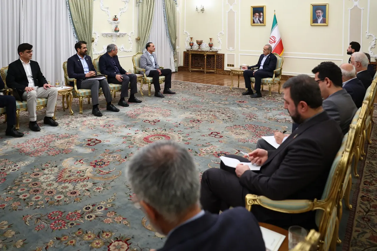 Diplomatic meeting in ornate room; delegations seated across carpet, Iranian flag and portraits visible.