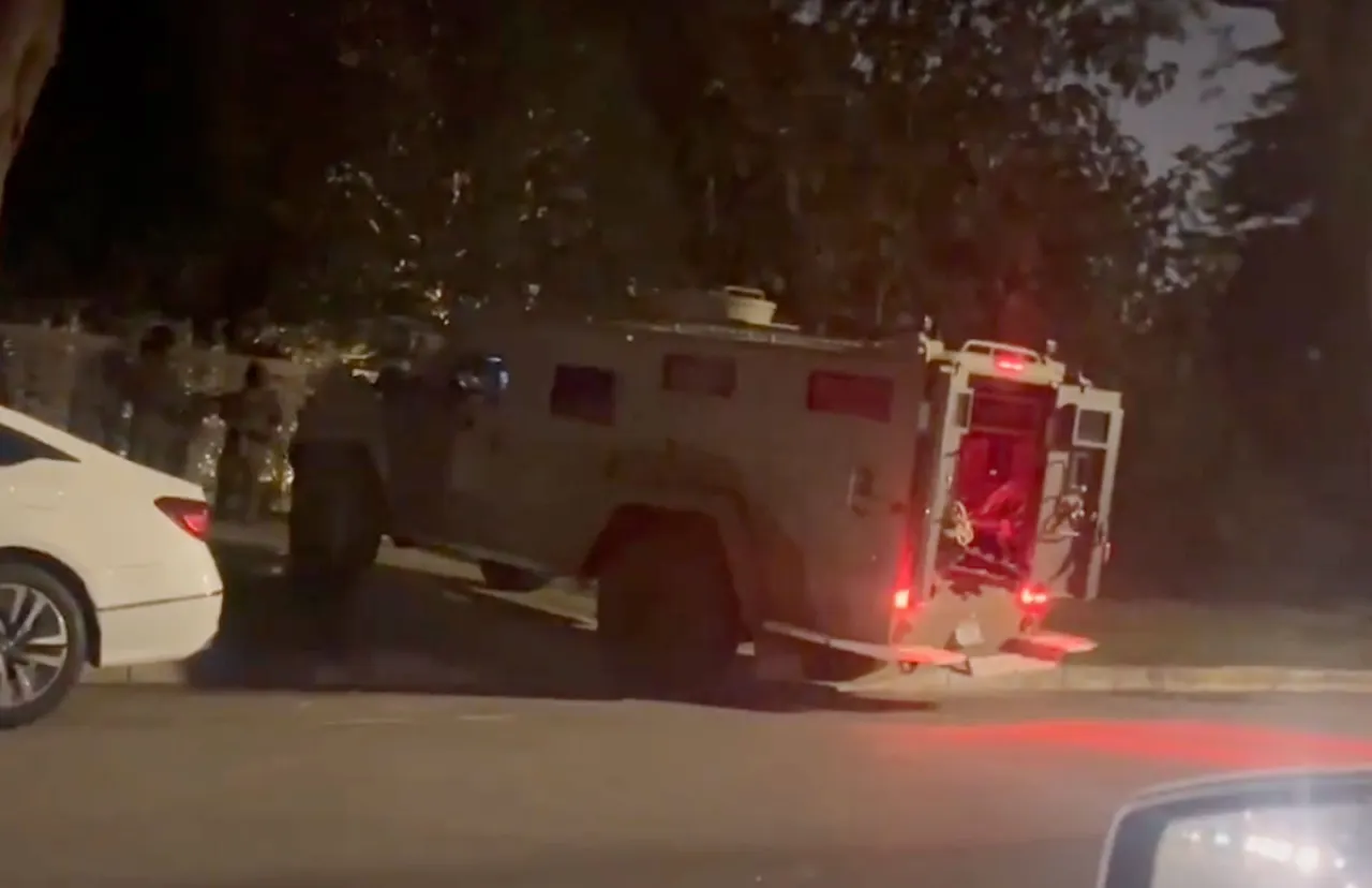 Armored tactical vehicle parked at night on a residential street beside a white car.