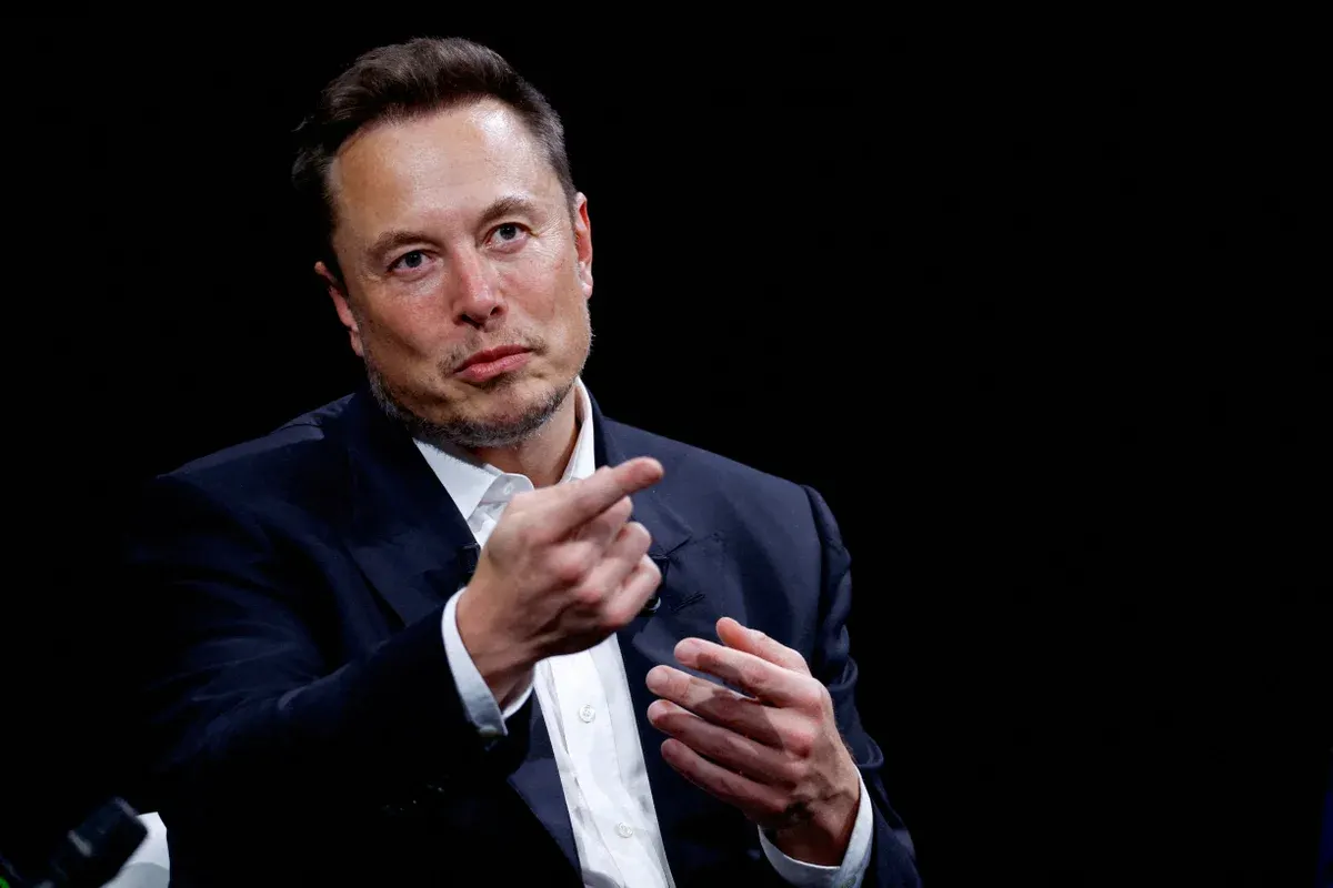 Man in dark suit and open collar gesturing while speaking against a black background.