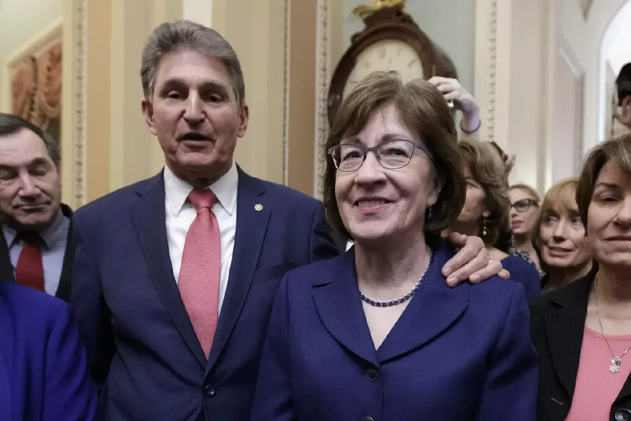 Two smiling politicians in dark suits posing indoors in a formal hallway with others behind
